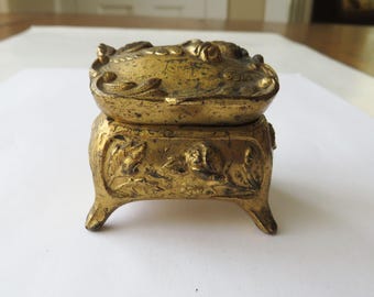 May include: A small, ornate, gold-colored metal trinket box with a hinged lid and intricate floral designs. The box has four legs and is decorated with raised floral motifs.