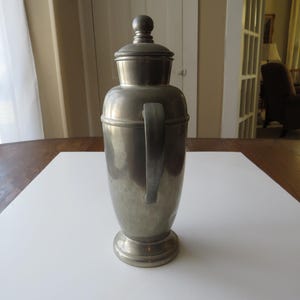 May include: A vintage silver-toned cocktail shaker with a handle and a lid. The shaker has a rounded body and a pedestal base. The handle is attached to the side of the shaker. The lid has a small knob on top. The shaker is sitting on a white surface.