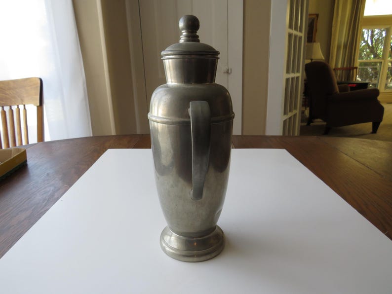 May include: A vintage silver-toned cocktail shaker with a handle and a lid. The shaker has a rounded body and a pedestal base. The handle is attached to the side of the shaker. The lid has a small knob on top. The shaker is sitting on a white surface.