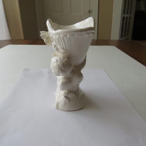 K&ouml;nnte beinhalten: Eine wei&szlig;e Keramikvase mit geriffeltem, offenem Oberteil, getragen von einem skulptierten Sockel mit cherubartigen Figuren. Die Vase hat ein klassisches, elegantes Design, geeignet f&uuml;r Blumengestecke oder dekorative Zwecke. Die Vase ist etwa 20 cm hoch.