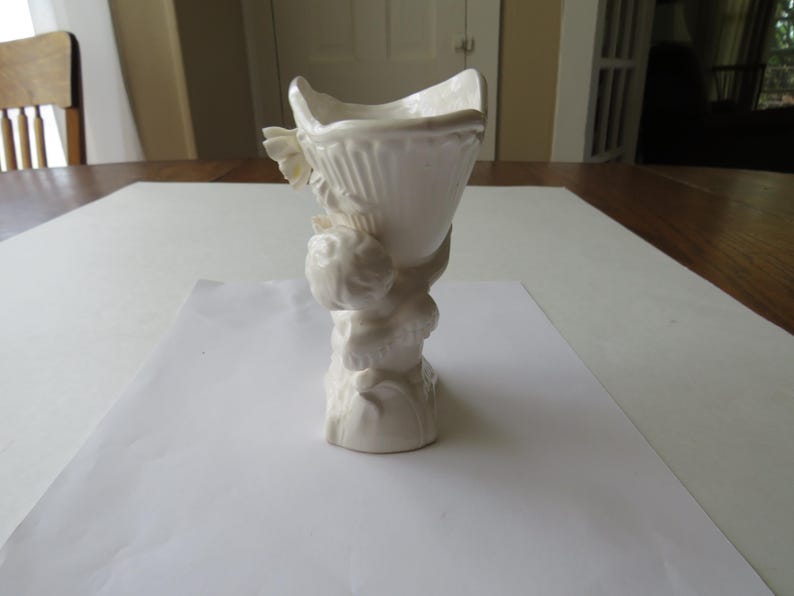 K&ouml;nnte beinhalten: Eine wei&szlig;e Keramikvase mit geriffeltem, offenem Oberteil, getragen von einem skulptierten Sockel mit cherubartigen Figuren. Die Vase hat ein klassisches, elegantes Design, geeignet f&uuml;r Blumengestecke oder dekorative Zwecke. Die Vase ist etwa 20 cm hoch.