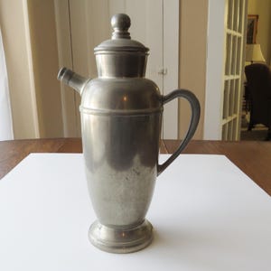 May include: A vintage, silver-toned cocktail shaker with a lid and a spout. The shaker has a handle and a rounded base. The metal surface shows signs of age and use. The shaker is designed for mixing drinks.
