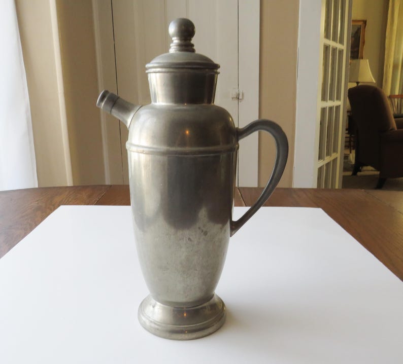 May include: A vintage, silver-toned cocktail shaker with a lid and a spout. The shaker has a handle and a rounded base. The metal surface shows signs of age and use. The shaker is designed for mixing drinks.