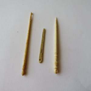 May include: Three antique-style crafting tools in a light cream color. The set includes a long crochet hook with a textured handle, a flat, narrow needle with a small eye, and a pointed stylus featuring a decorative, grooved handle design.