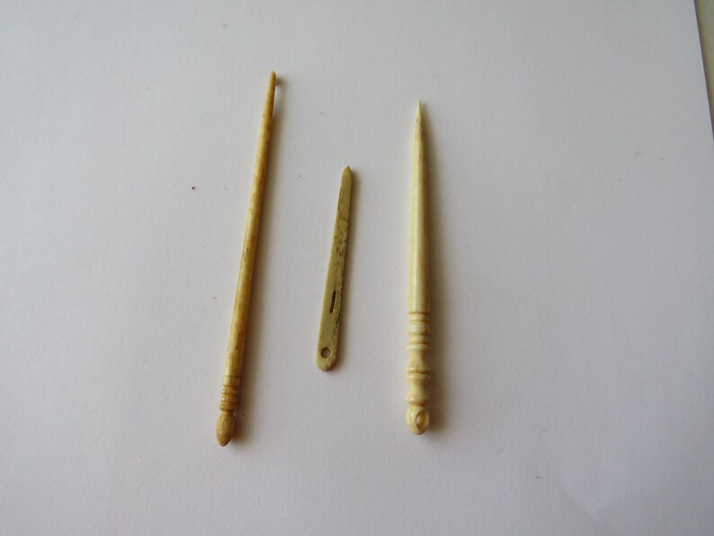 May include: Three antique-style crafting tools in a light cream color. The set includes a long crochet hook with a textured handle, a flat, narrow needle with a small eye, and a pointed stylus featuring a decorative, grooved handle design.