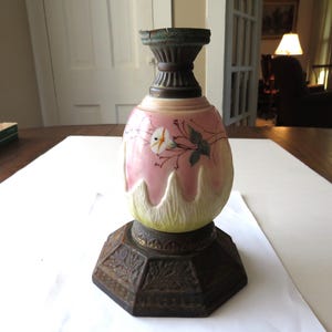 May include: A glass lamp base featuring a pink and cream gradient body with a hand-painted white flower and green leaves. The base has a dark, textured metal pedestal with an octagonal shape and a matching dark metal collar at the top opening.