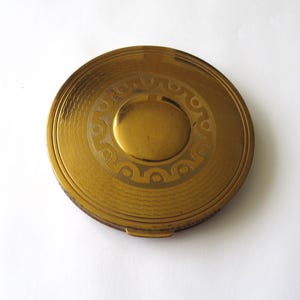 May include: A round, gold-toned metal compact case featuring a smooth, polished center circle surrounded by a textured band with a repeating geometric wave and circle pattern. The outer edge of the lid has a fine, wavy line engraving.