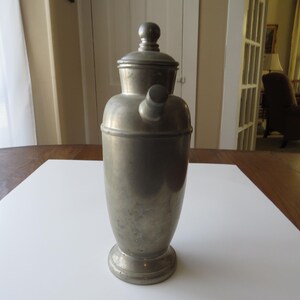 May include: A vintage silver-toned cocktail shaker with a rounded body, a lid, and a spout. The shaker is made of metal and sits on a round base. It is displayed on a white surface on a wooden table.