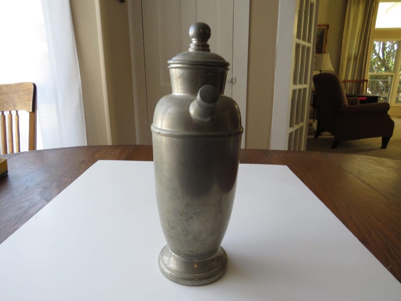 May include: A vintage silver-toned cocktail shaker with a rounded body, a lid, and a spout. The shaker is made of metal and sits on a round base. It is displayed on a white surface on a wooden table.