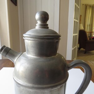 May include: A vintage metal coffee pot with a lid and a handle. The pot is silver-colored and has a spout for pouring. The design is simple and classic, suggesting a traditional style. The pot is likely made of pewter or a similar metal.