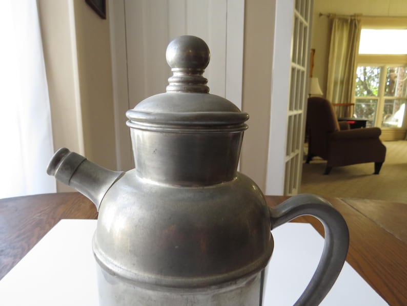 May include: A vintage metal coffee pot with a lid and a handle. The pot is silver-colored and has a spout for pouring. The design is simple and classic, suggesting a traditional style. The pot is likely made of pewter or a similar metal.