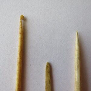 May include: Three light beige, textured tools with a mottled appearance. One tool features a hooked end for crocheting, while the other two are pointed, straight implements. The items have a smooth, organic finish and are arranged vertically.