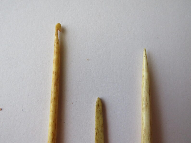 May include: Three light beige, textured tools with a mottled appearance. One tool features a hooked end for crocheting, while the other two are pointed, straight implements. The items have a smooth, organic finish and are arranged vertically.