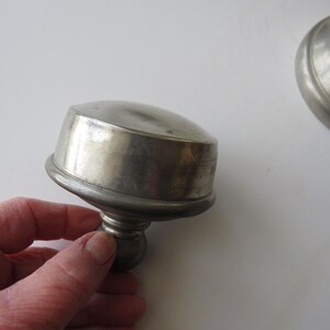 May include: A pewter-colored metal lid with a rounded top and a decorative base. The lid is slightly tarnished, showing signs of age. The image suggests a vintage or antique item, possibly a container lid.