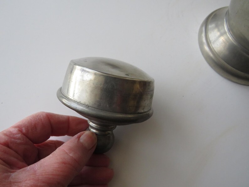 May include: A pewter-colored metal lid with a rounded top and a decorative base. The lid is slightly tarnished, showing signs of age. The image suggests a vintage or antique item, possibly a container lid.