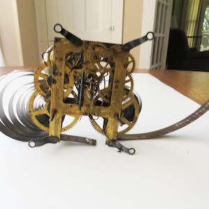 May include: A disassembled antique clock mechanism. The brass-colored gears and springs are arranged on a white surface. The clock's internal components are visible, including the intricate gear system and coiled springs.