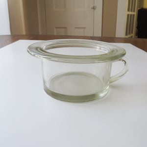 May include: A clear glass chamber pot with a handle. The pot has a wide, flat rim and a rounded base. The glass is slightly tinted, and the pot is sitting on a white surface.