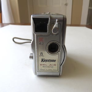 May include: A vintage Keystone Bel Air Magazine 8mm movie camera. The silver camera has a black lens and a gray strap. The text "K 42" and "8 mm" are visible on the front. The camera is marked "Keystone BEL AIR MAGAZINE" and "BOSTON 24, MASS. U.S.A."