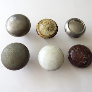 May include: A collection of six round cabinet knobs arranged in two rows. The set includes a brushed metal knob, a weathered textured knob, a smooth silver-toned knob, a dark matte knob, a white ceramic knob, and a glossy dark brown knob.