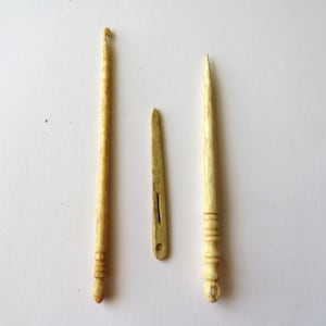 May include: Three antique-style crafting tools made of a light beige, textured material. The set includes a crochet hook with a curved tip, a flat needle with a central slit and circular eye, and a pointed tool featuring a decorative ringed handle.