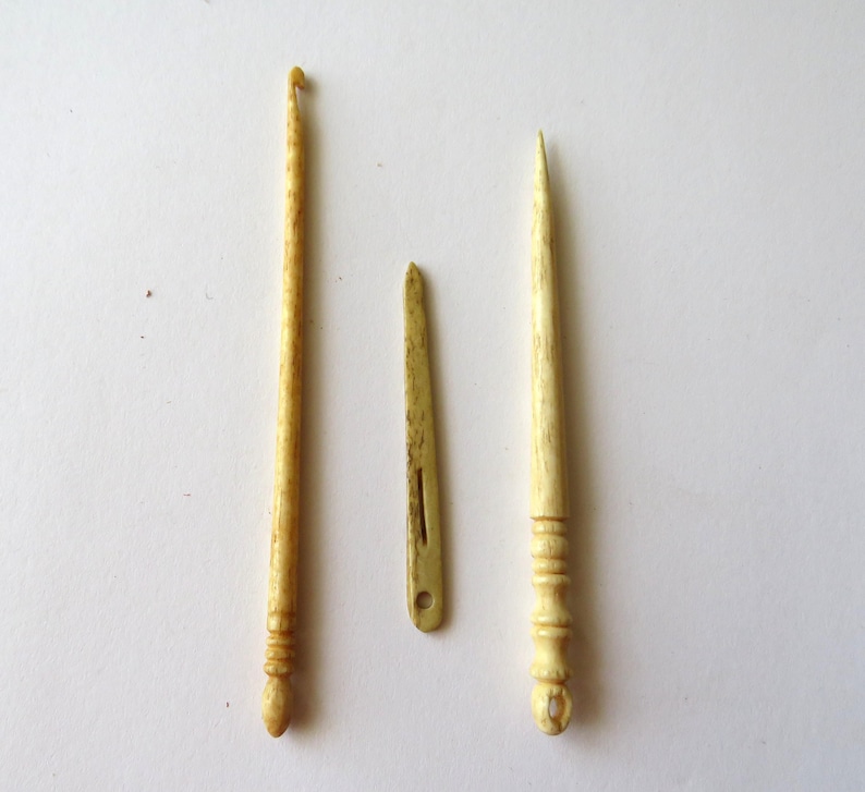 May include: Three antique-style crafting tools made of a light beige, textured material. The set includes a crochet hook with a curved tip, a flat needle with a central slit and circular eye, and a pointed tool featuring a decorative ringed handle.