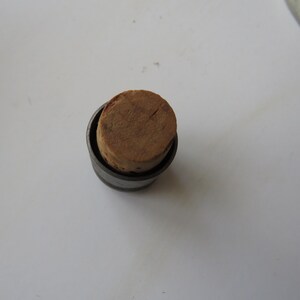 May include: A small, cylindrical bottle stopper with a natural cork top. The cork is a light brown color and fits snugly inside a dark metal casing. The stopper is designed to seal a bottle.