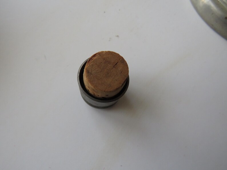 May include: A small, cylindrical bottle stopper with a natural cork top. The cork is a light brown color and fits snugly inside a dark metal casing. The stopper is designed to seal a bottle.