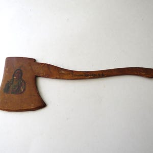 May include: A vintage wooden axe with a brown finish. The axe head features a painted image of a Native American figure. The curved handle has the text "Siloam Springs Ark."