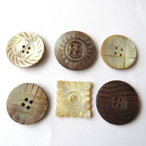 May include: A collection of six decorative buttons in various shapes and colors. The buttons include round and square designs, with colors ranging from light beige to dark brown. Some buttons have carved patterns, while others have a smooth finish.