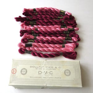 May include: A collection of twisted embroidery floss skeins in shades of pink and deep magenta, each secured with a black paper band. Below the floss sits a rectangular off-white cardboard box labeled Dollfus-Mieg & C, D.M.C, and Mulhouse-Belfort-Paris.