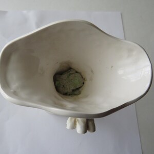 K&ouml;nnte beinhalten: Eine wei&szlig;e Keramikvase mit einer organischen, asymmetrischen Form. Die Vase hat einen breiten, ausgestellten Rand und ein strukturiertes Inneres. Im Inneren der Vase ist ein gr&uuml;ner, steinartiger Gegenstand zu sehen. Die Vase steht auf einer wei&szlig;en Oberfl&auml;che.