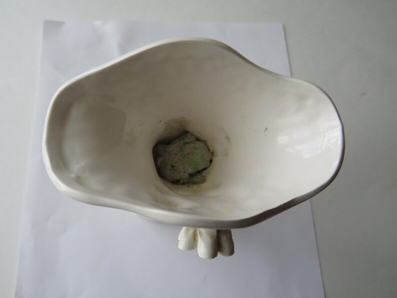 K&ouml;nnte beinhalten: Eine wei&szlig;e Keramikvase mit einer organischen, asymmetrischen Form. Die Vase hat einen breiten, ausgestellten Rand und ein strukturiertes Inneres. Im Inneren der Vase ist ein gr&uuml;ner, steinartiger Gegenstand zu sehen. Die Vase steht auf einer wei&szlig;en Oberfl&auml;che.