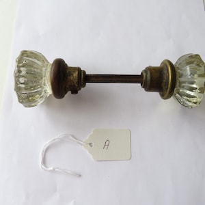 May include: Antique glass doorknob set with a ribbed design. The knobs are clear, with brass and brown metal hardware. A white tag with the letter "A" is attached.