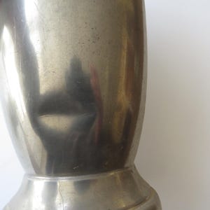 May include: A close-up of a vintage, silver-colored metal pitcher. The pitcher has a handle and a wide base. The surface of the pitcher has a slightly textured appearance with some visible wear and tear.