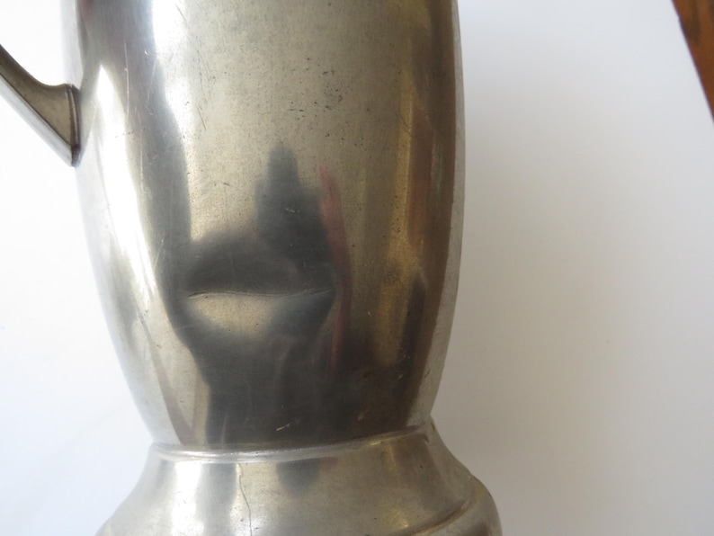 May include: A close-up of a vintage, silver-colored metal pitcher. The pitcher has a handle and a wide base. The surface of the pitcher has a slightly textured appearance with some visible wear and tear.