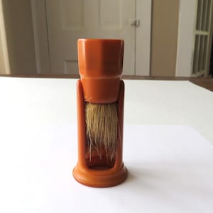 May include: An orange shaving brush with a cylindrical handle and a base. The brush bristles are light brown. The brush is standing upright on a white surface.