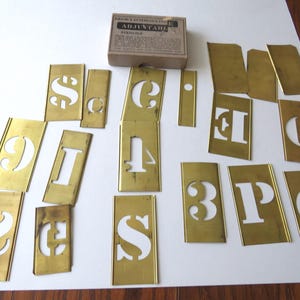 May include: A set of brass interlocking adjustable stencils featuring various letters, numbers, and symbols, accompanied by a cardboard box labeled 'Reese's Interlocking Adjustable Stencils'. The individual metal plates have cutouts for characters.