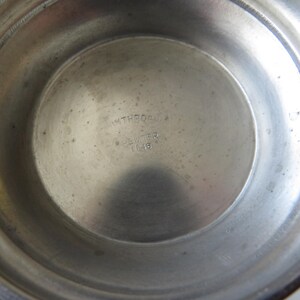 May include: A close-up of the interior of a pewter bowl. The bowl has a circular base with the text "INTREQS, PEWTER, 1146" stamped in the center. The bowl's surface is a dull silver color with minor imperfections.