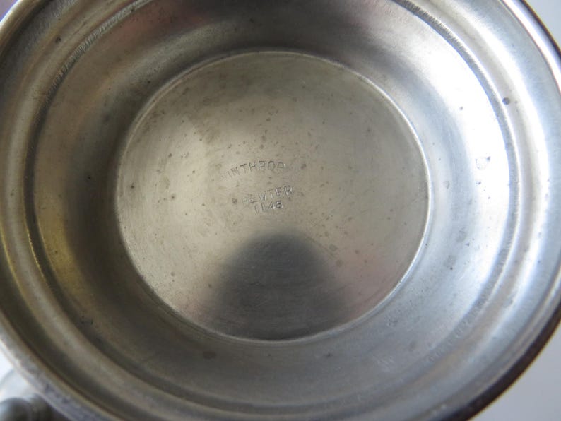 May include: A close-up of the interior of a pewter bowl. The bowl has a circular base with the text "INTREQS, PEWTER, 1146" stamped in the center. The bowl's surface is a dull silver color with minor imperfections.