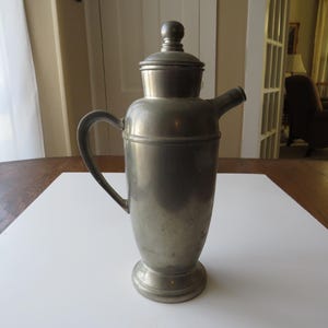 May include: A vintage metal cocktail shaker with a handle, spout, and lid. The shaker is silver-toned and sits on a white surface. The design is classic and elegant, suitable for home bars or vintage decor.