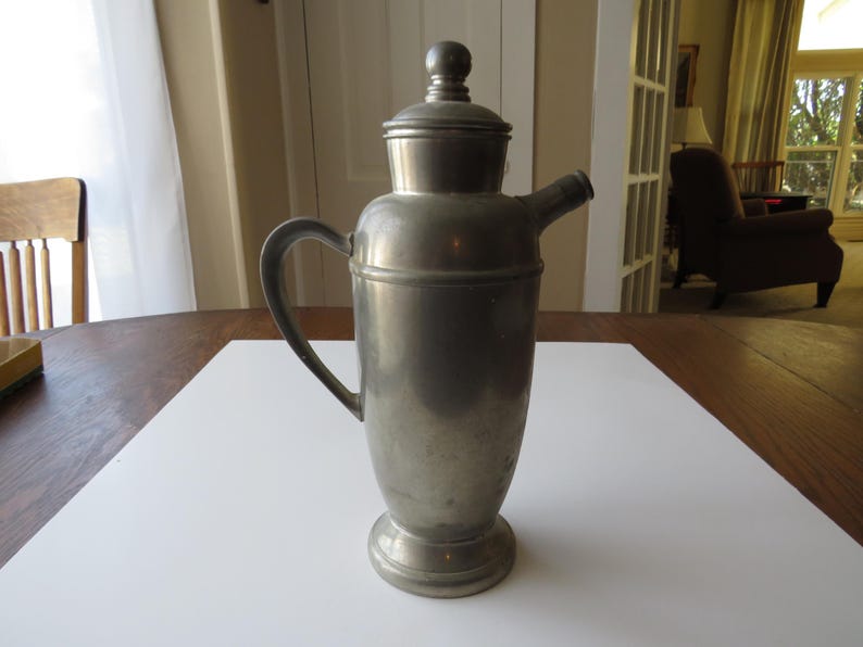 May include: A vintage metal cocktail shaker with a handle, spout, and lid. The shaker is silver-toned and sits on a white surface. The design is classic and elegant, suitable for home bars or vintage decor.