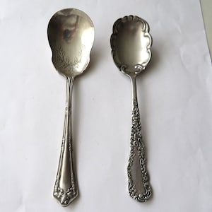 May include: Two antique silver spoons. One spoon has a wide, oval bowl with a floral design. The other spoon has a scalloped bowl and ornate handle. Both spoons are displayed on a white surface.