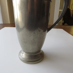 May include: A vintage silver-colored metal pitcher with a handle. The pitcher has a wide base and a slightly tapered body. The handle is curved and attached to the side of the pitcher. The pitcher is sitting on a white surface.