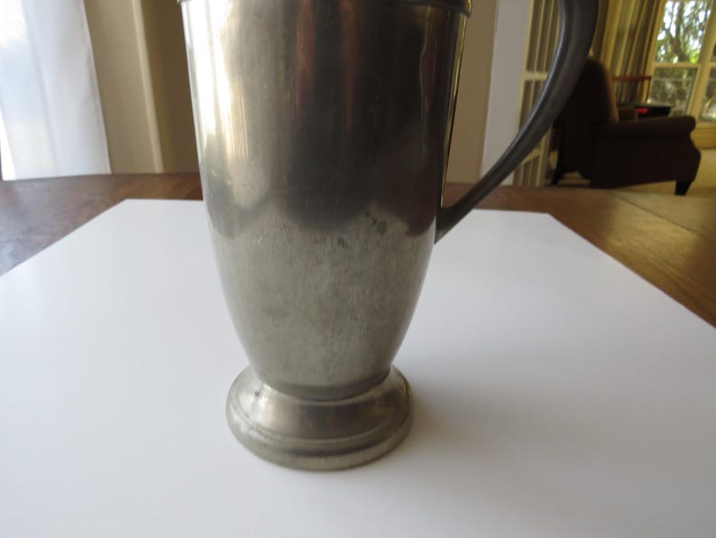 May include: A vintage silver-colored metal pitcher with a handle. The pitcher has a wide base and a slightly tapered body. The handle is curved and attached to the side of the pitcher. The pitcher is sitting on a white surface.
