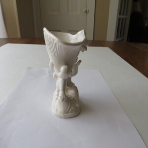 K&ouml;nnte beinhalten: Eine wei&szlig;e Keramikvase mit einzigartigem Design. Die Vase hat eine ausgestellte Oberseite mit einem strukturierten, gerippten Muster und wird von einem geformten Sockel mit Figuren getragen. Die Gesamth&ouml;he betr&auml;gt ca. 18 cm.