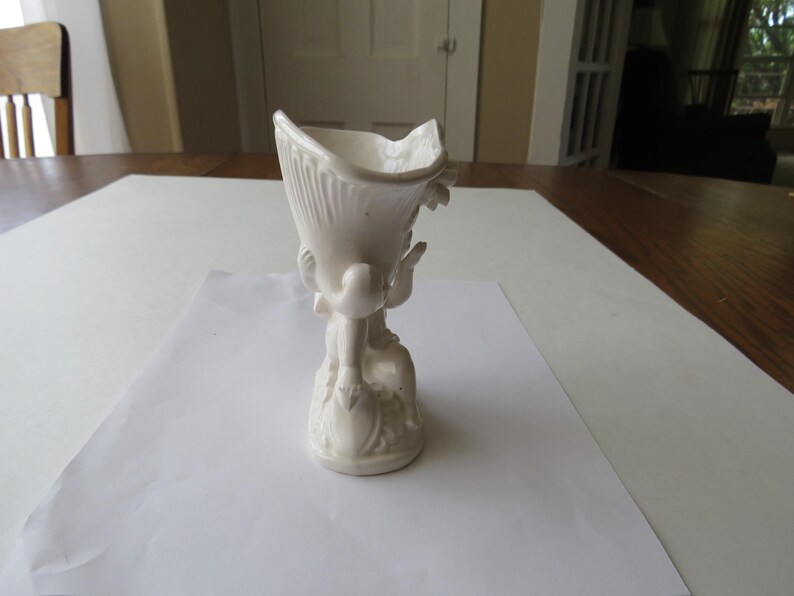 K&ouml;nnte beinhalten: Eine wei&szlig;e Keramikvase mit einzigartigem Design. Die Vase hat eine ausgestellte Oberseite mit einem strukturierten, gerippten Muster und wird von einem geformten Sockel mit Figuren getragen. Die Gesamth&ouml;he betr&auml;gt ca. 18 cm.