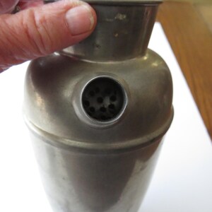 May include: A vintage metal cocktail shaker with a rounded body and a conical top. The shaker has a small strainer opening and a tarnished, aged appearance. The image shows the shaker being held by a person.