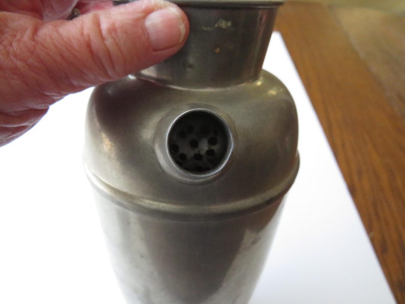May include: A vintage metal cocktail shaker with a rounded body and a conical top. The shaker has a small strainer opening and a tarnished, aged appearance. The image shows the shaker being held by a person.