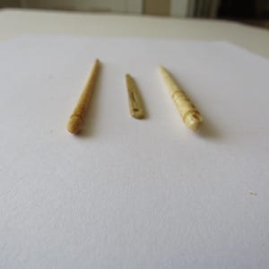 May include: Three small, light beige, carved objects arranged in a row. The outer two items are slender, pointed tools with rounded, textured ends. The center item is a flat, thin, elongated piece with a small hole near one end.