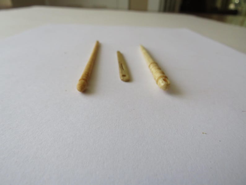 May include: Three small, light beige, carved objects arranged in a row. The outer two items are slender, pointed tools with rounded, textured ends. The center item is a flat, thin, elongated piece with a small hole near one end.